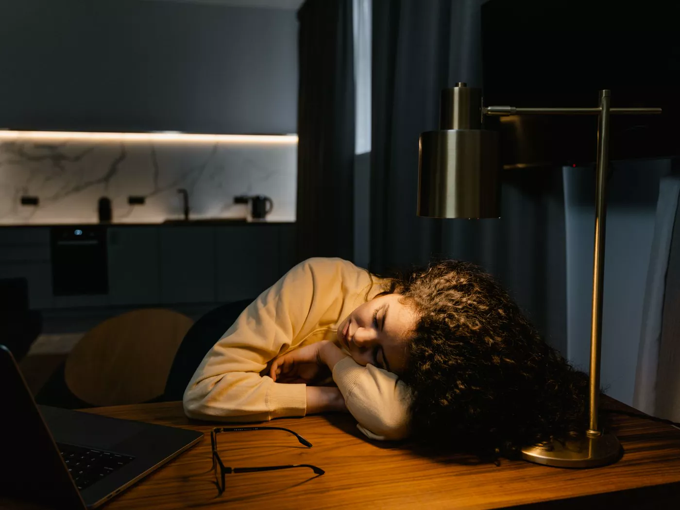A tired woman with curly hair asleep at a desk with a laptop and glasses under warm lamp light in a dimly lit room.