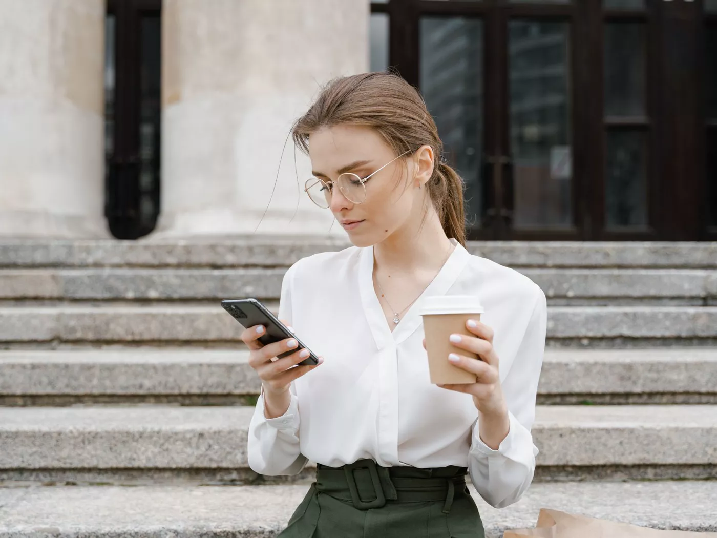 Young woman in business attire using smartphone while enjoying a coffee outdoors.