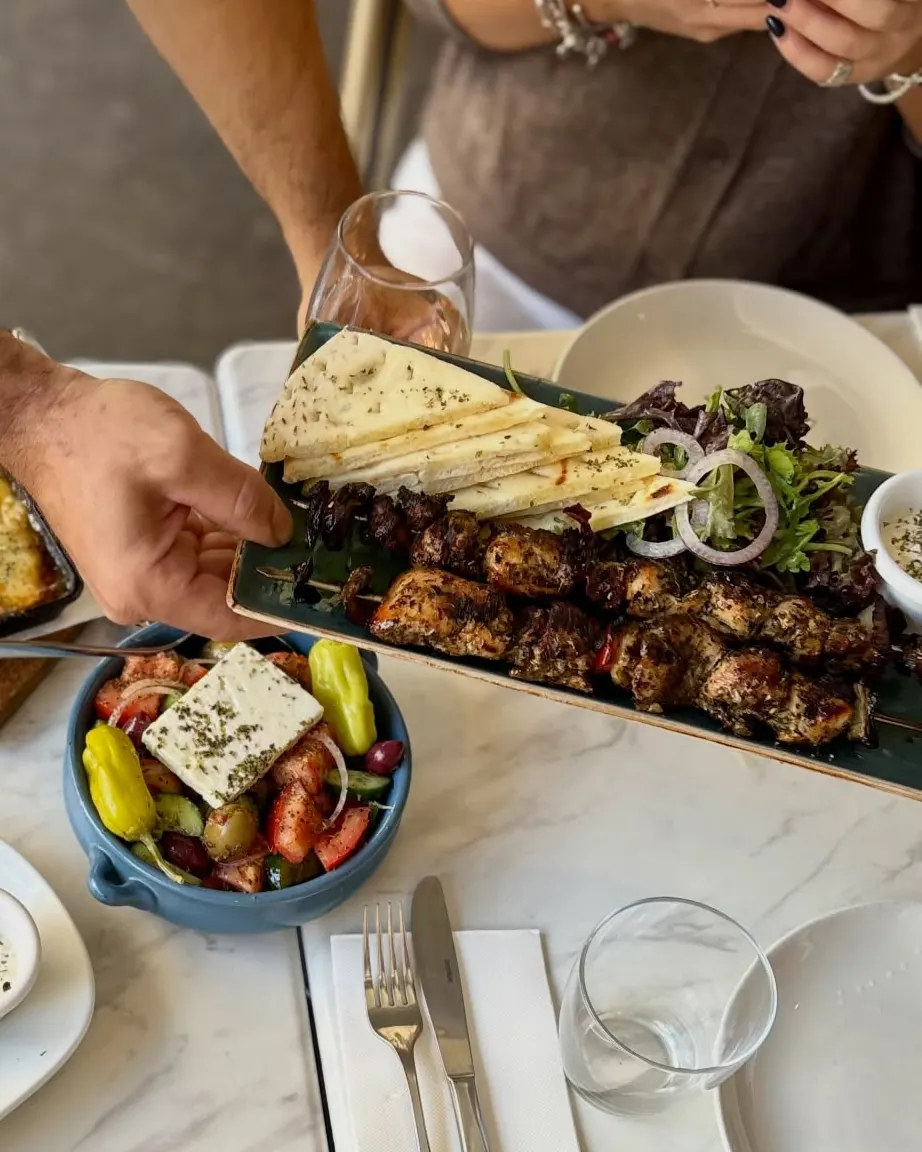 A platter of chargrilled souvlaki skewers with grilled pita and salad being served, beside a Greek salad
