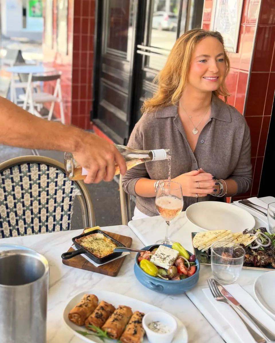 Wine poured for a smiling guest at a footpath table beside the red-tiled shopfront, with Greek share plates on the marble table