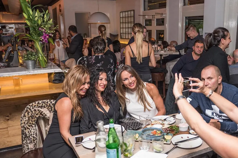 Friends posing for a photo at a table of Greek share plates on a packed Greek Night at The Phoenix, with diners and the lit bar behind