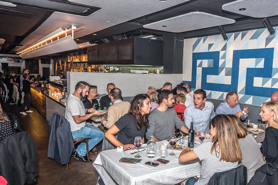 Guests at long tables under the blue and white Greek-key mural during a Phoenix feast