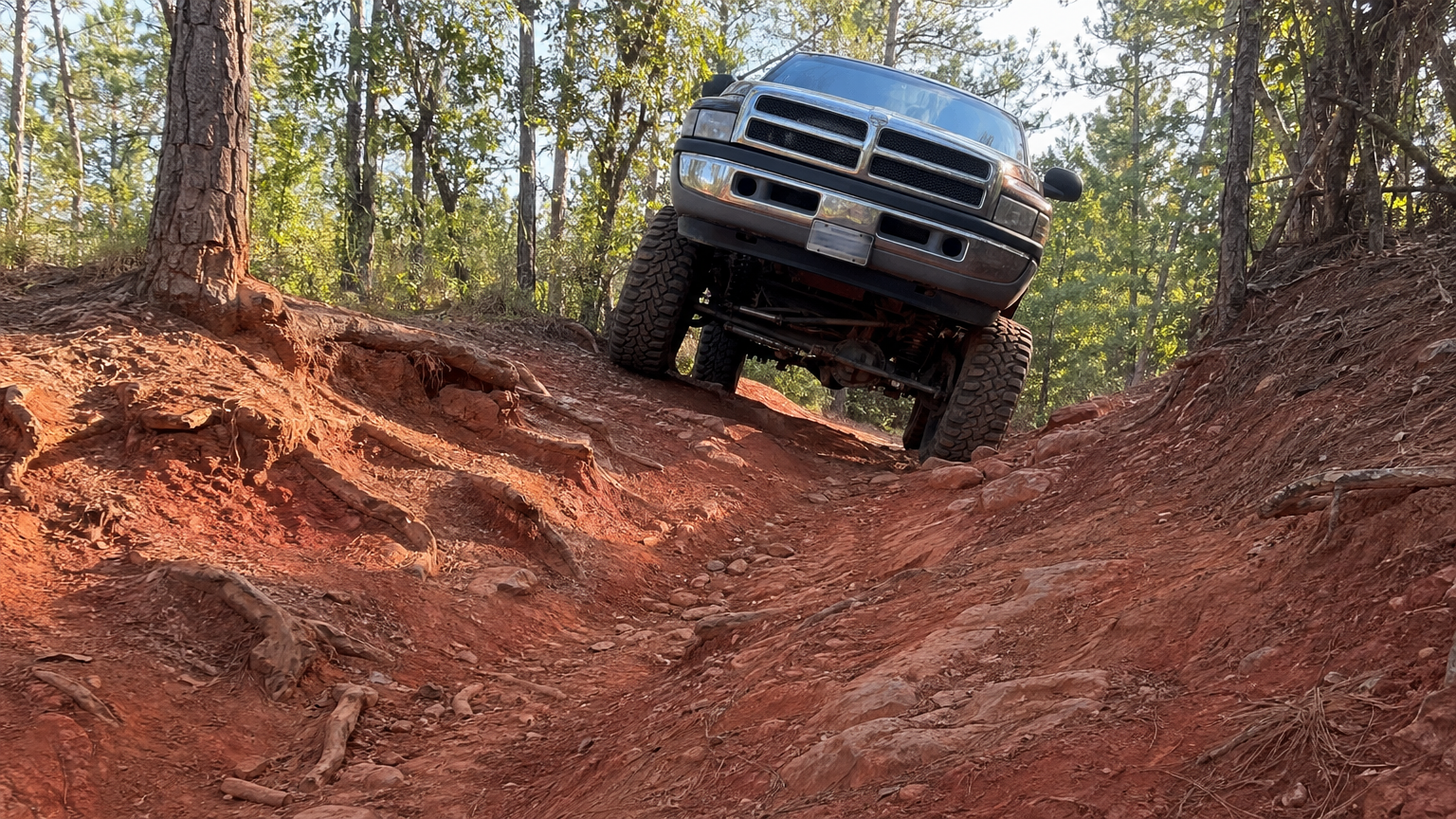 Lifted truck navigating steep off-camber red dirt hill climb through East Texas pine forest