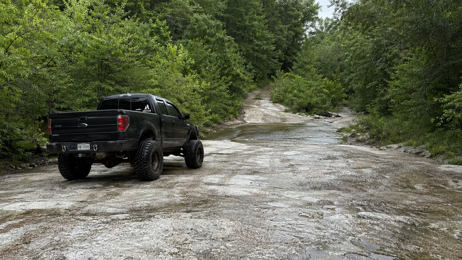 Lifted truck on slick Louisiana limestone outcrop with water crossing visible ahead