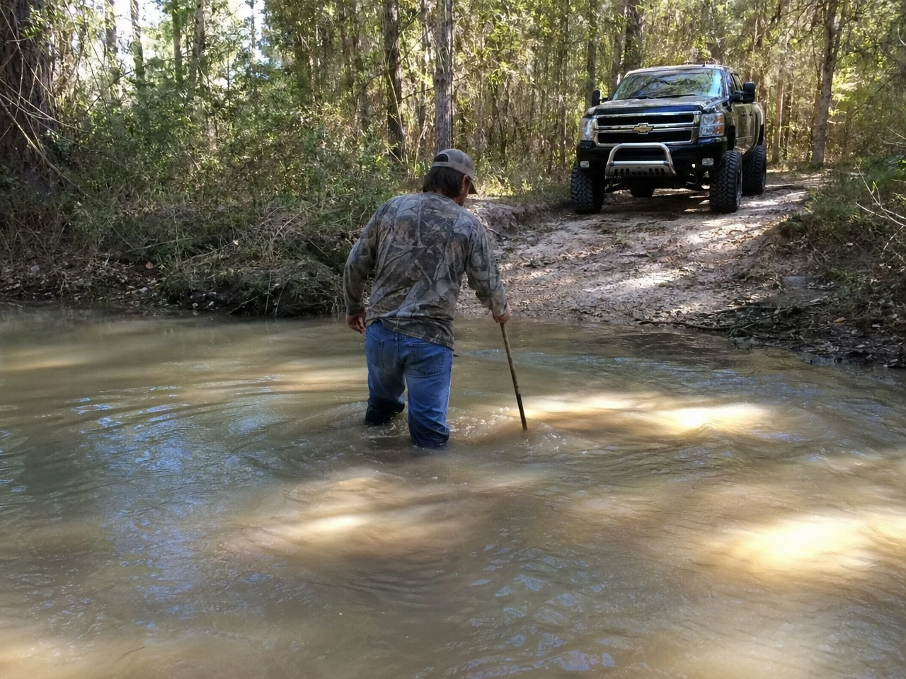 Person wading Texas creek crossing knee-deep assessing depth with lifted truck waiting on bank