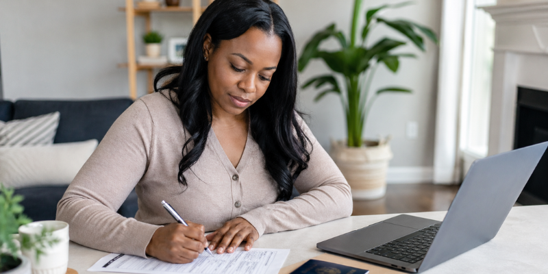 An African American woman sits at a clean desk in a bright home, calmly filling out a U.S. passport application with her ID, documents, and laptop nearby