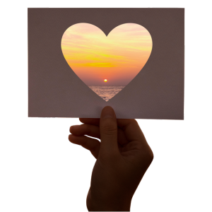 Photo of a hand holding a postcard sized card with a heart shape cut out of it and a gorgeous sunset is visible through the heart-shaped hole.