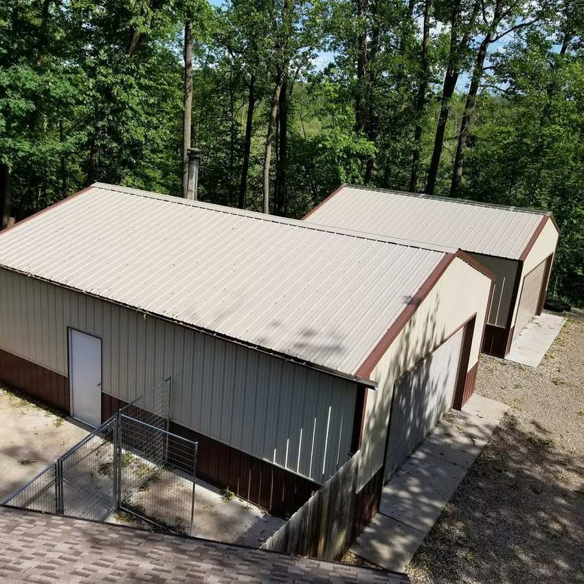 Two tan steel pole buildings with metal roofs among tall trees on an acreage in Hugo, MN, seen from the house roof during a Repair King storm inspection