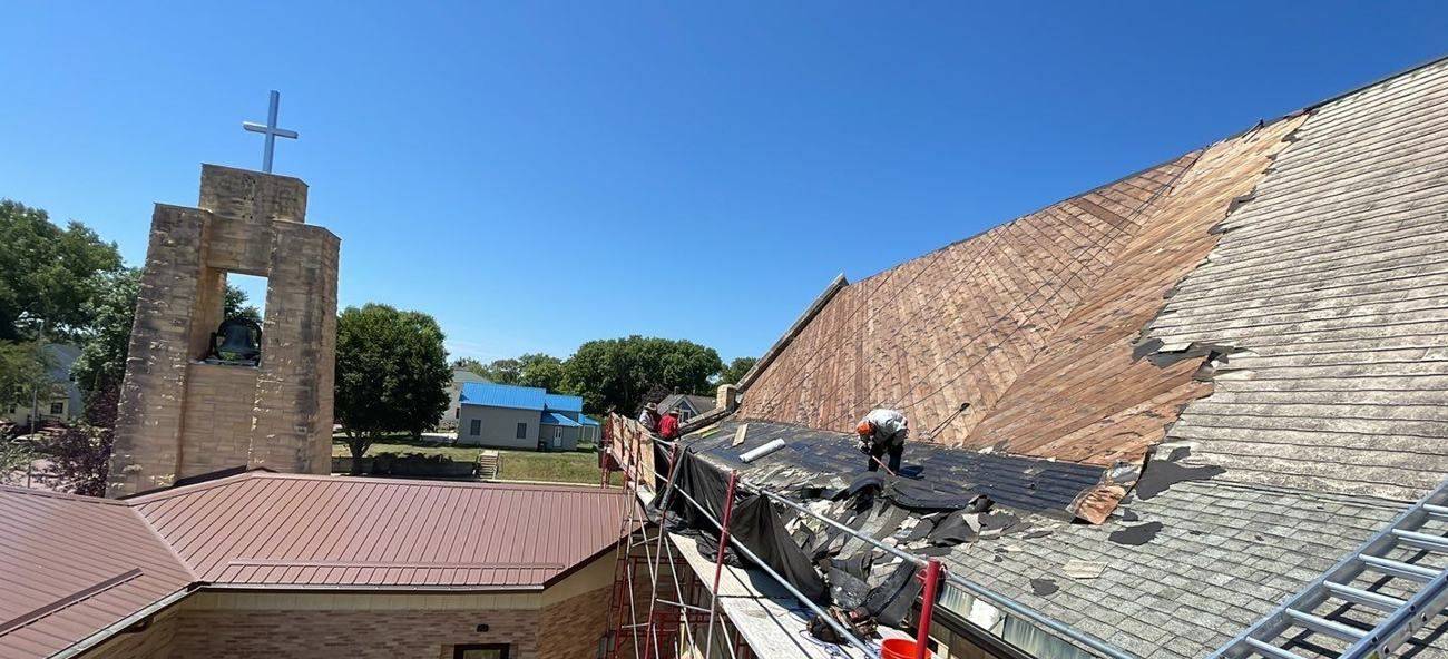 Repair King roofers tearing off the steep roof of St. Paul's Lutheran Church in St. James, MN, beside the stone bell tower and cross, with scaffolding and guardrails along the eave, July 2023