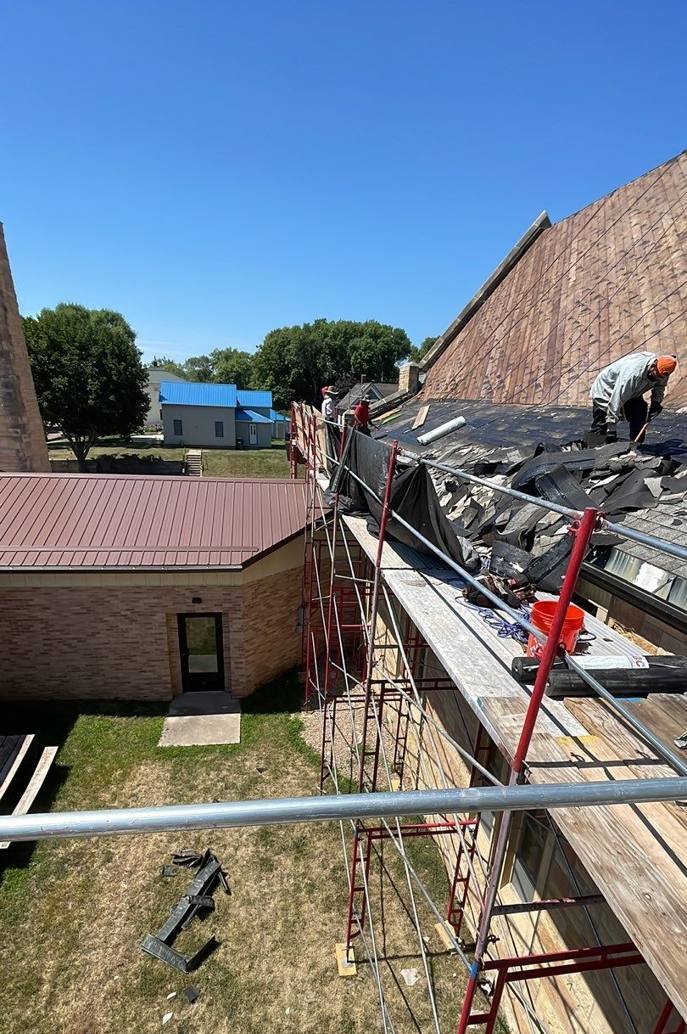 Repair King crew tearing off the steep roof of St. Paul's Lutheran Church in St. James, MN, from scaffolding beside the red metal wing roof, July 2023