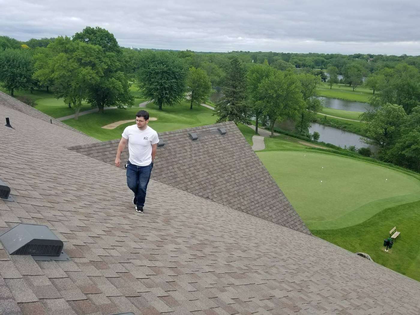Repair King inspector walking the shingle roof of the Owatonna Country Club clubhouse in Owatonna, MN, above a putting green, ponds and the tree-lined golf course