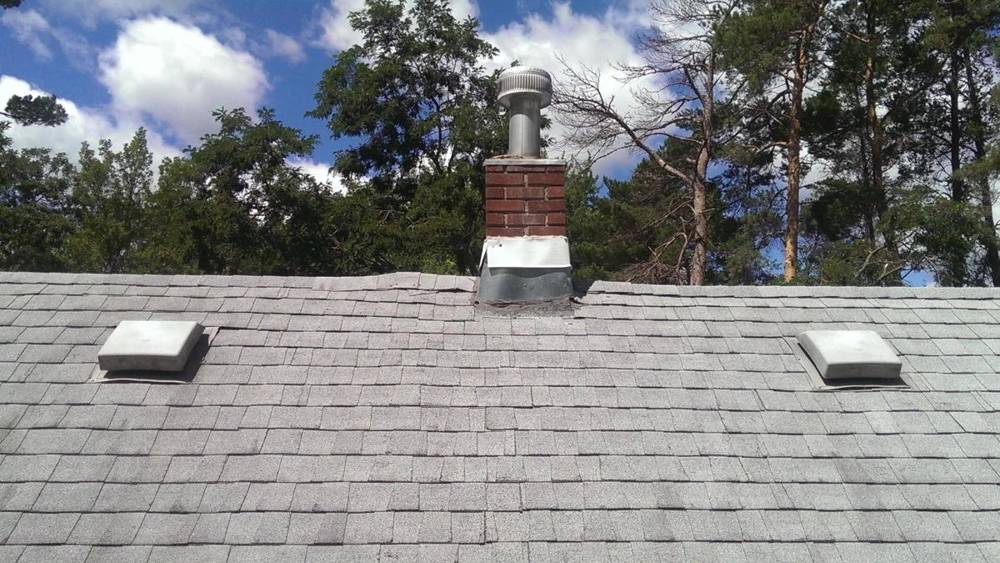 Weathered gray asphalt shingles, box vents and a brick chimney on a Hinckley home before Repair King's multi-layer tear-off, with tall pines behind