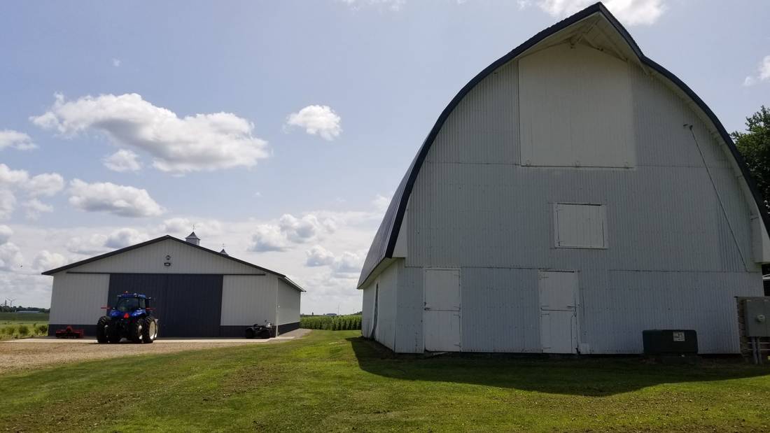 White barn with a gambrel-style arched roof beside a steel machine shed and a tractor on a farm near Blue Earth, MN