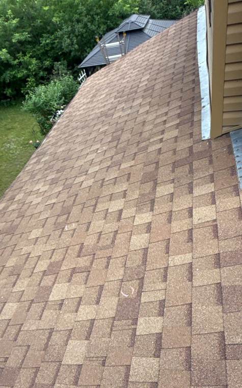 Brown shingle roof slope running along tan siding toward the trees on a wooded Stacy, MN lot, June 2026