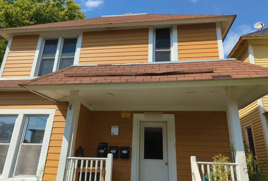 Two-story lap-sided rental house with three mailboxes on the porch in the Elliot Park neighborhood of Minneapolis, with a loose strip of shingles on the porch roof, August 2023