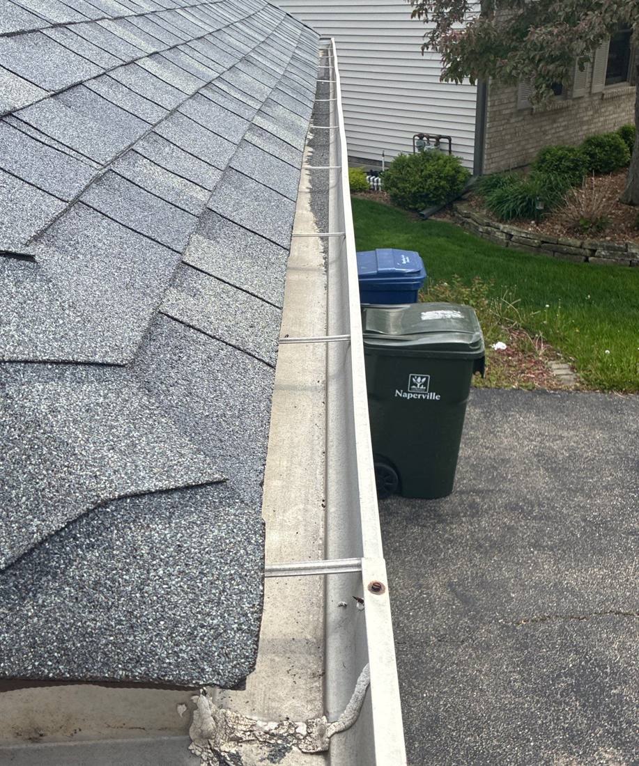 Gutter and roof edge on a Naperville home, with the city's recycling and trash bins in the driveway below, May 2026
