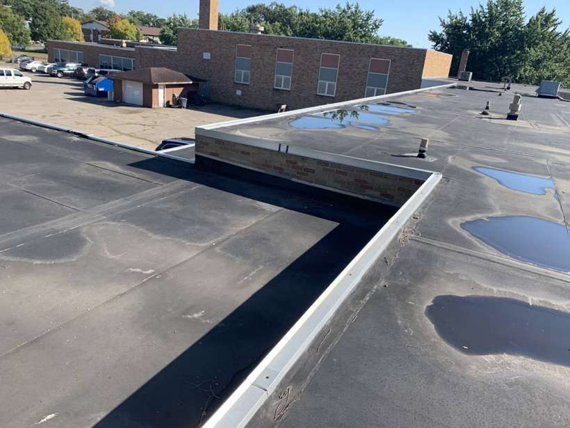 Ponded water on the multi-level black rubber roof at Washington Place in St. Cloud, MN, with the brick building behind, during a Repair King inspection