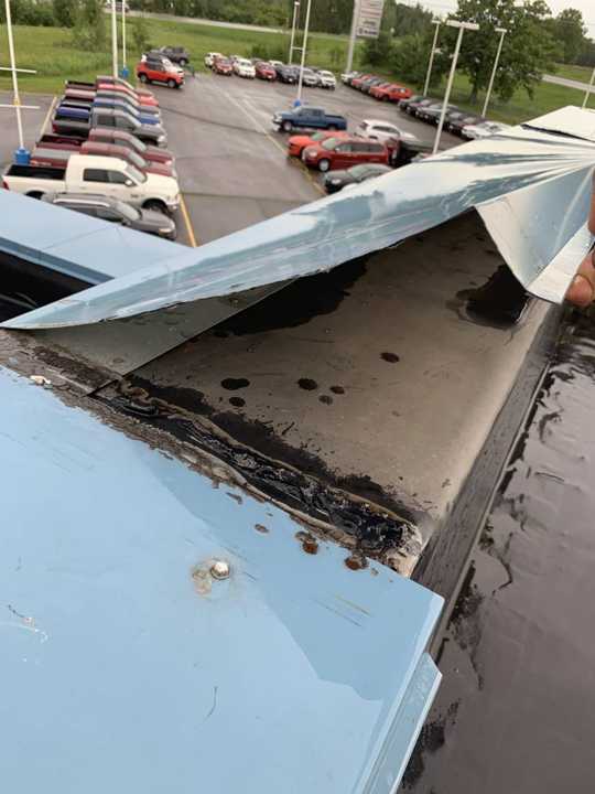 A lifted section of blue metal coping at the edge of the Eide Chrysler roof in Pine City, MN, with the dealership lot below, July 2019