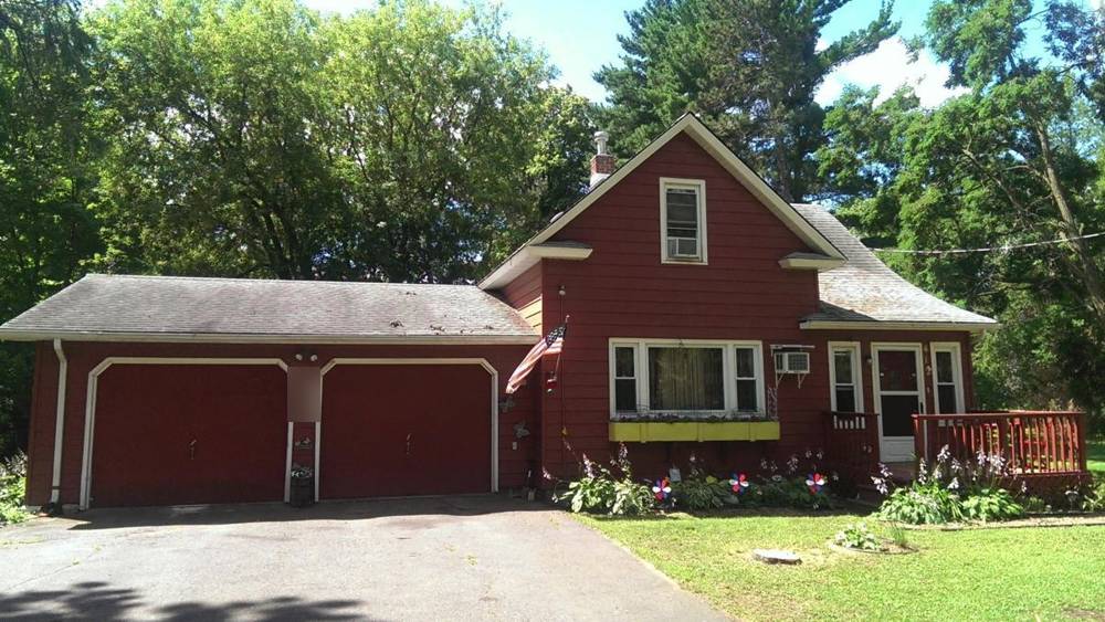The Hinckley home before the work: red siding, weathered shingle roof, brick chimney and attached two-car garage under tall pines, summer