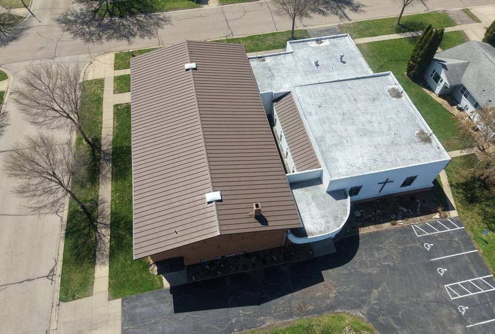 Aerial view of Bethany Lutheran Church in Lake City, MN: brown metal sanctuary roof beside the white flat membrane roofs and rounded entry canopy, April 2026