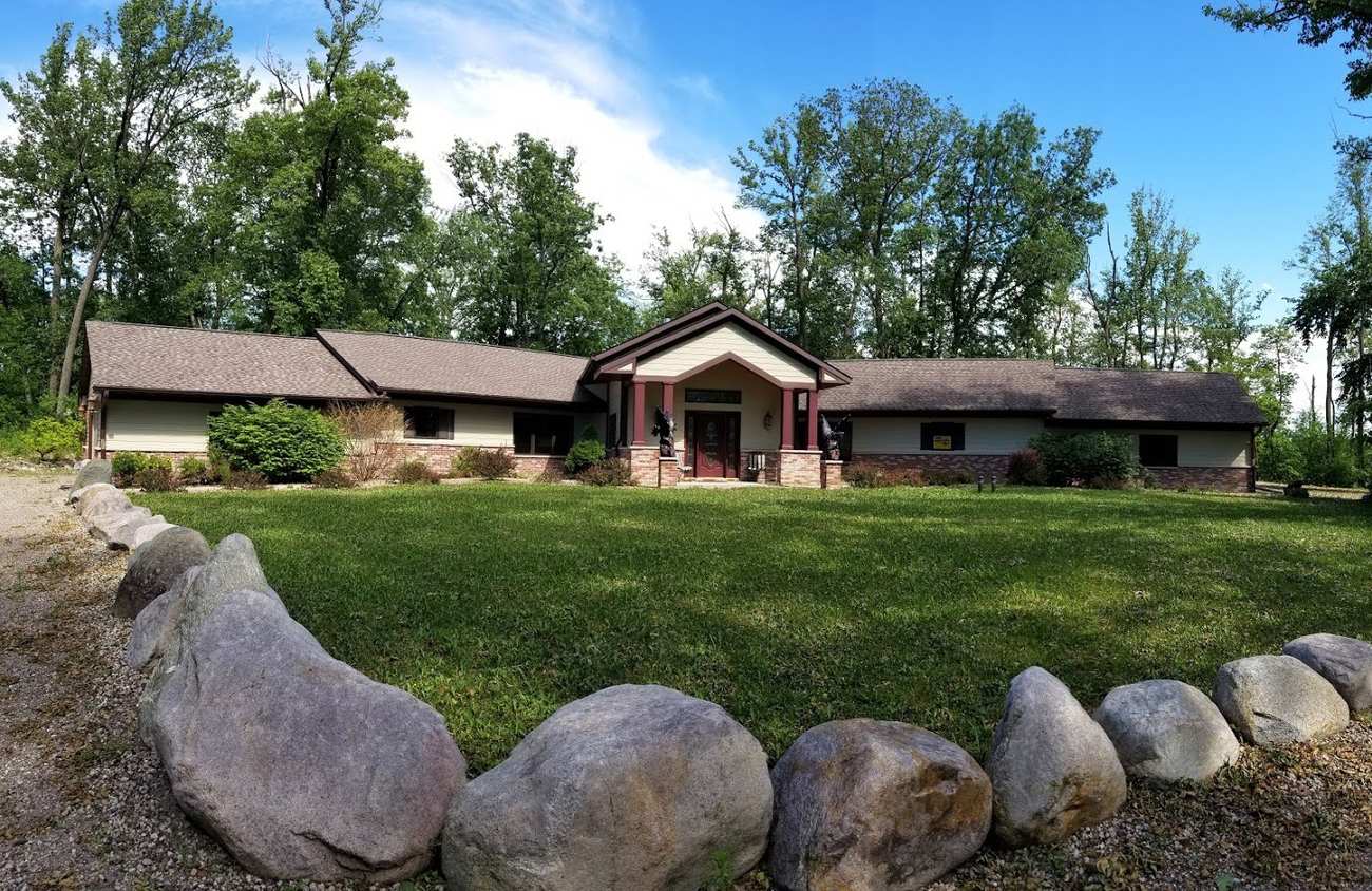 Wooded ranch-style acreage home in Hugo, MN with a brown shingle roof, brick wainscot and a boulder-lined lawn, documented by Repair King