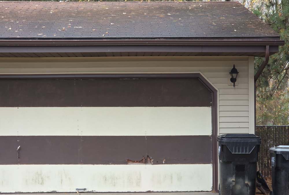 Garage of a vinyl-sided rambler in Columbus, MN, with an aging brown shingle roof and a weathered two-tone garage door, photographed by Repair King in October 2025