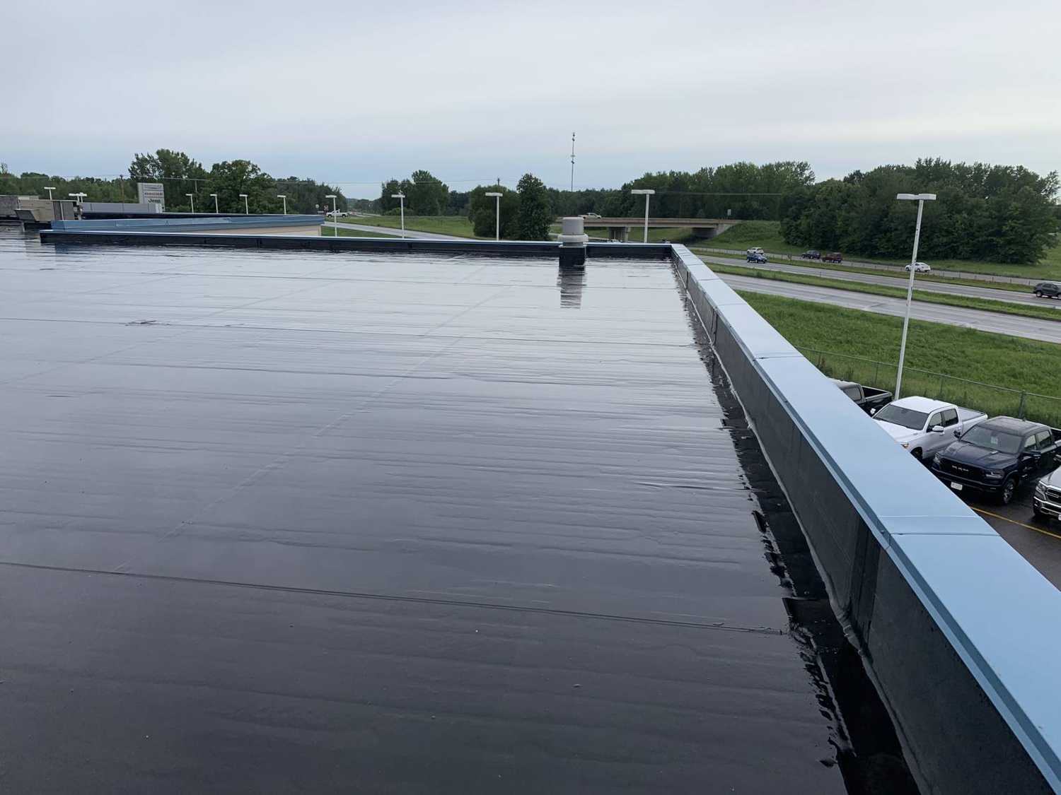The black rubber flat roof and metal coping at Eide Chrysler Dodge Jeep Ram in Pine City, MN, with the lot and I-35 behind it, photographed by Repair King on July 20, 2019