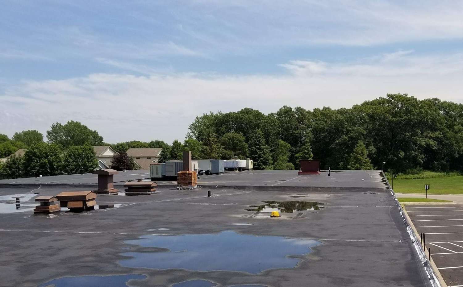 Standing water across the flat rubber roof of King of Glory Lutheran Church in Blaine, MN, with trees and the parking lot beyond, July 2017