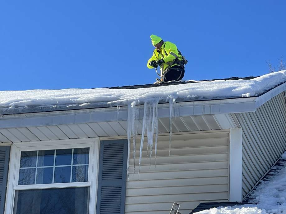 Harnessed crew member in high-visibility gear working an ice dam on a roof