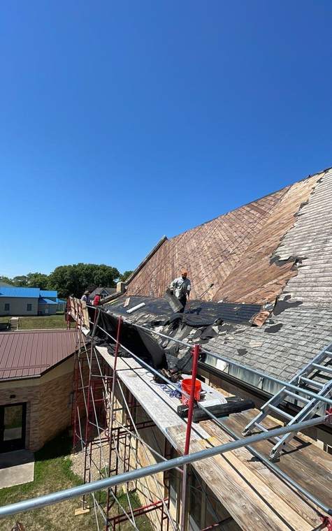 Repair King roofer tearing off old shingles on the steep roof of St. Paul's Lutheran Church in St. James, MN, working from scaffolding with a red guardrail