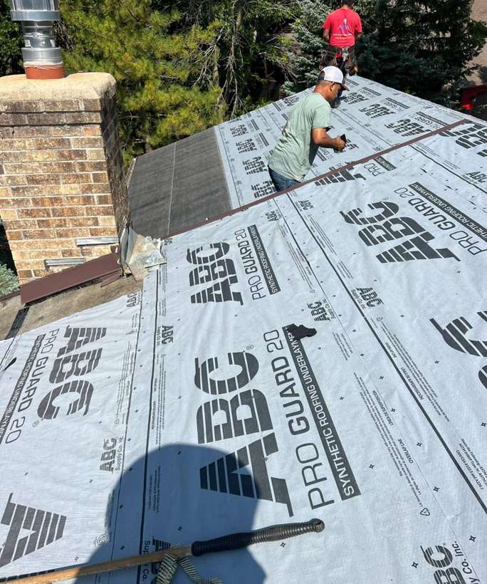 Crew installing synthetic roofing underlayment beside a brick chimney on a Lakeville home, September 2023