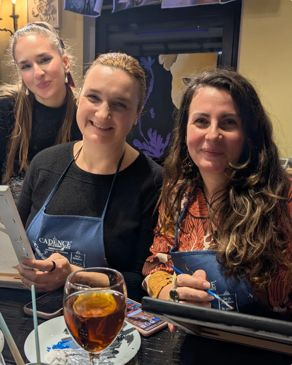 Trei cursante zâmbind la masa din atelier, cu pensulele în mână și un pahar de rosé