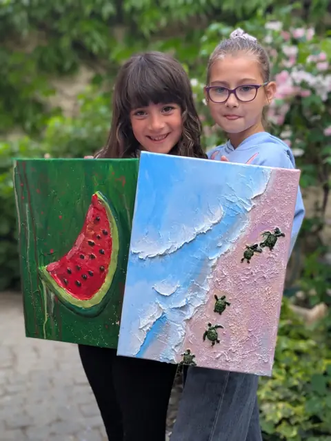 Două fetițe cu primele lor tablouri în relief: o felie de pepene și țestoase pe plajă
