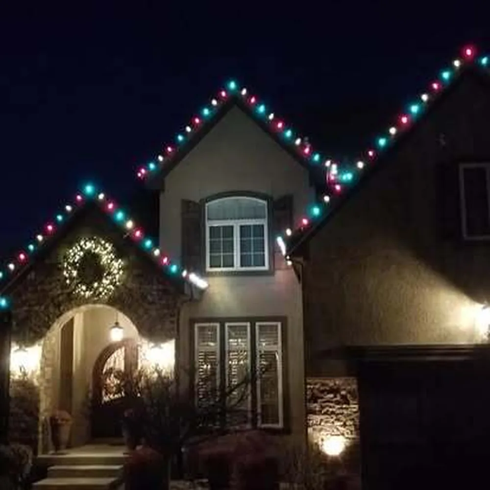 Teal, pink and white C9 bulbs on a two-story stone home with a wreath over the entry