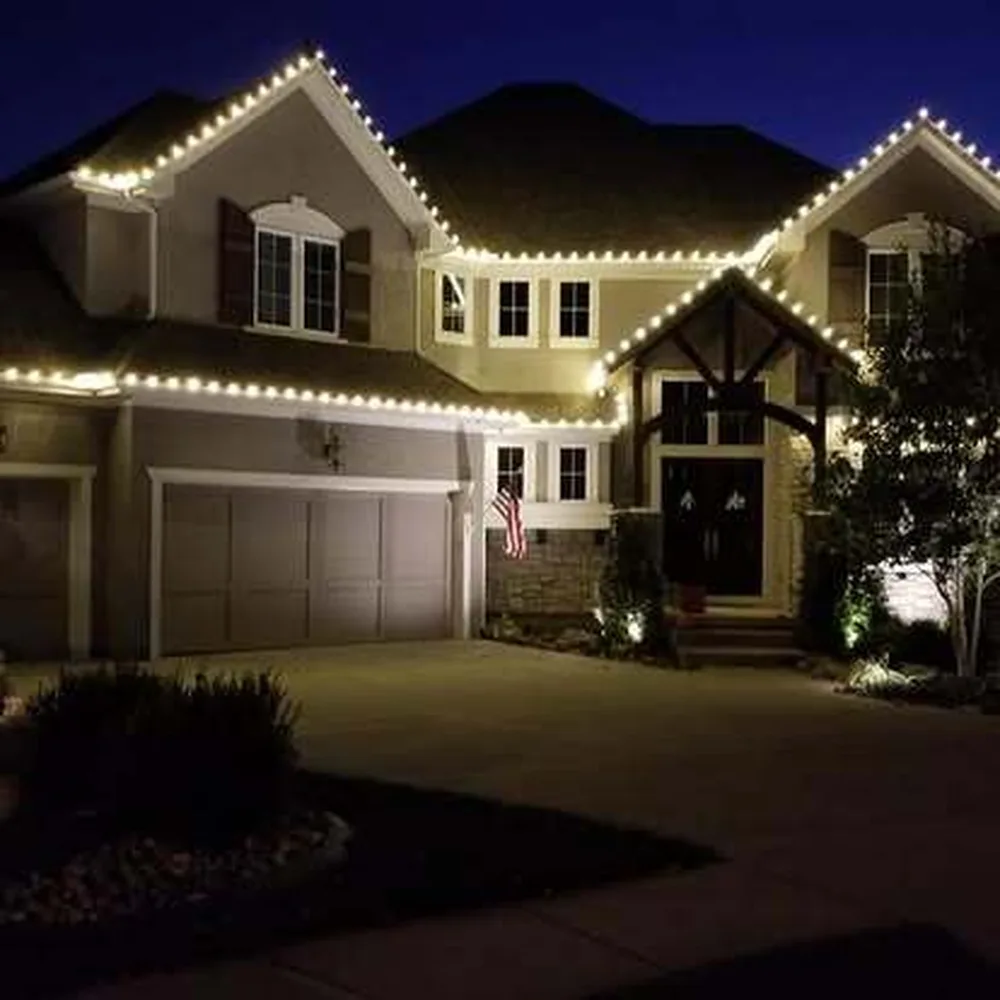 Warm white C9 bulbs tracing the roofline of a tan two-story home with an American flag by the entry