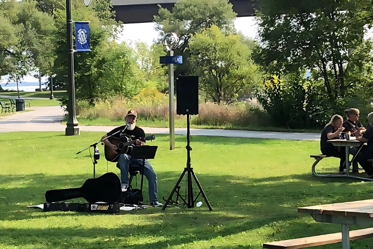 Solo musician performing acoustic guitar near picnic tables with guests enjoying lunch at Pop-Ups on the Bay in West Zwick’s Park, Belleville.
