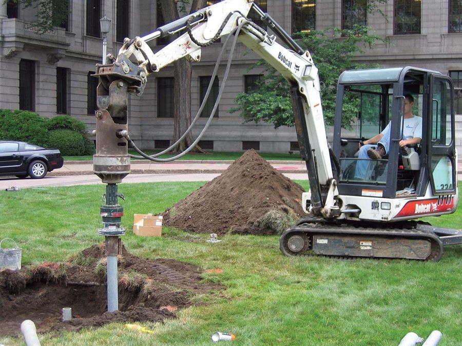 Compact excavator with a drive head installing a helical pile in front of a large building