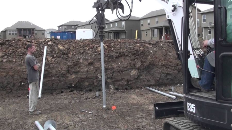 Mini excavator driving a helical pile at a residential development while a crew member stands nearby