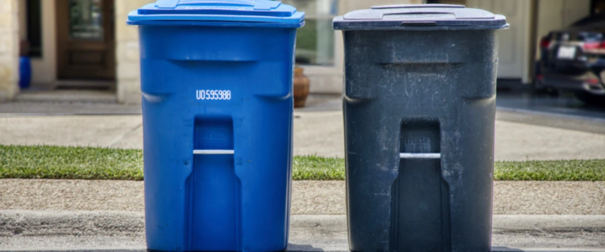 Blue recycling bin and black trash bin placed curbside in a residential neighborhood. Blue recycling bin and black trash bin placed curbside in a residential neighborhood.