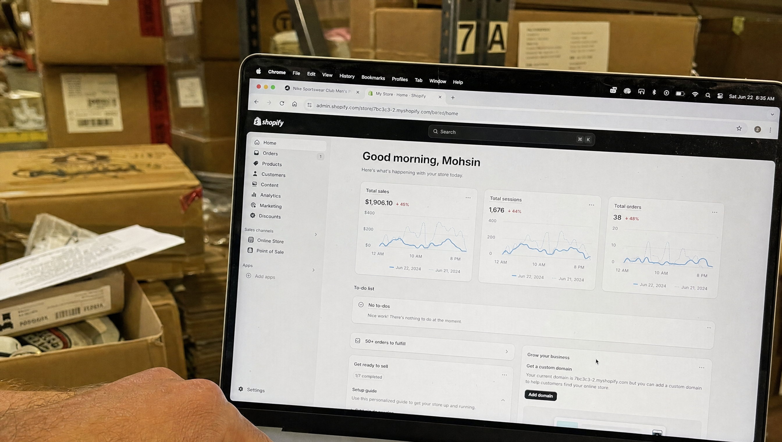 Laptop showing a Shopify store dashboard, warehouse boxes stacked in the background