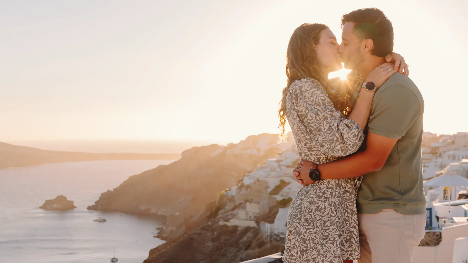 Elise and Ricardo kissing on a whitewashed dome in Oia, Santorini, the sun setting over the caldera behind them