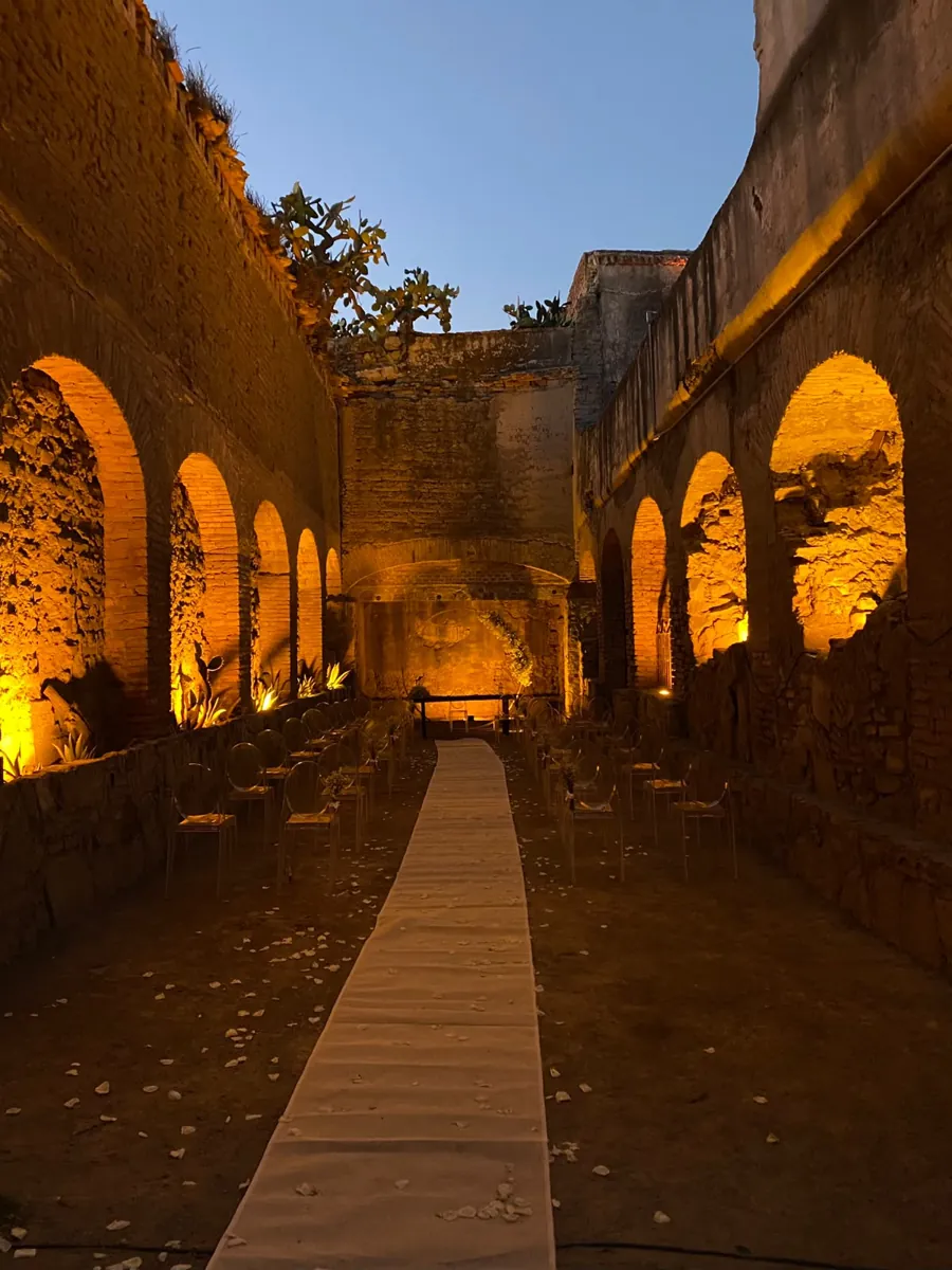 The roofless nave of Hacienda Concepci&oacute;n at night, every arch lit amber, chairs lining the aisle