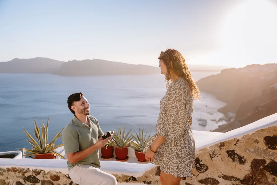 Ricardo looks at Elise across the terrace wall, the sea bright behind her