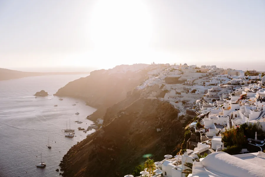 Oia from above at golden hour: the caldera, the whitewashed houses stepping down the cliff, boats in the bay below