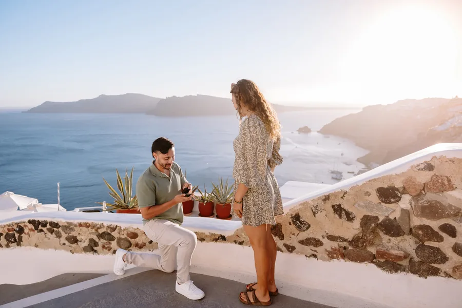 Ricardo sits on the terrace wall while Elise stands beside him, both looking out to sea