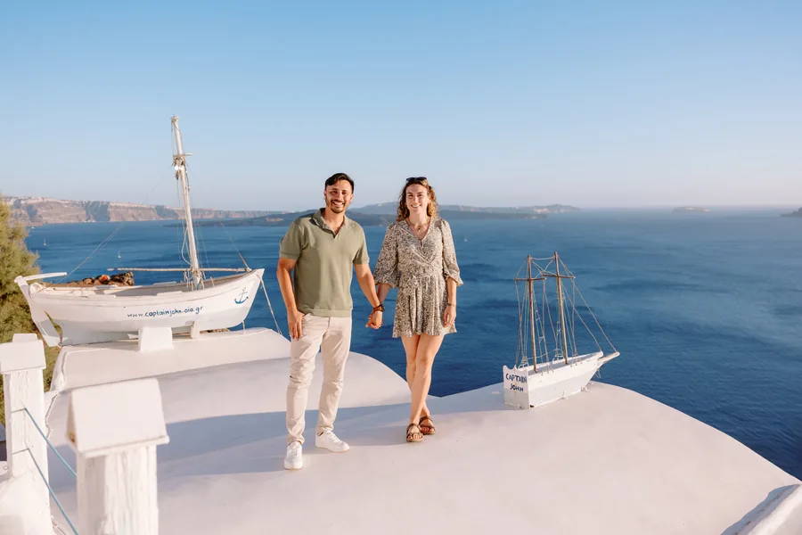 Elise and Ricardo walking across a white rooftop in Oia, a small boat parked on it, the caldera a deep blue behind