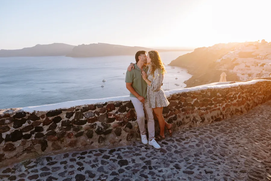 Ricardo’s arm around Elise at the stone wall, Oia’s cliffs behind