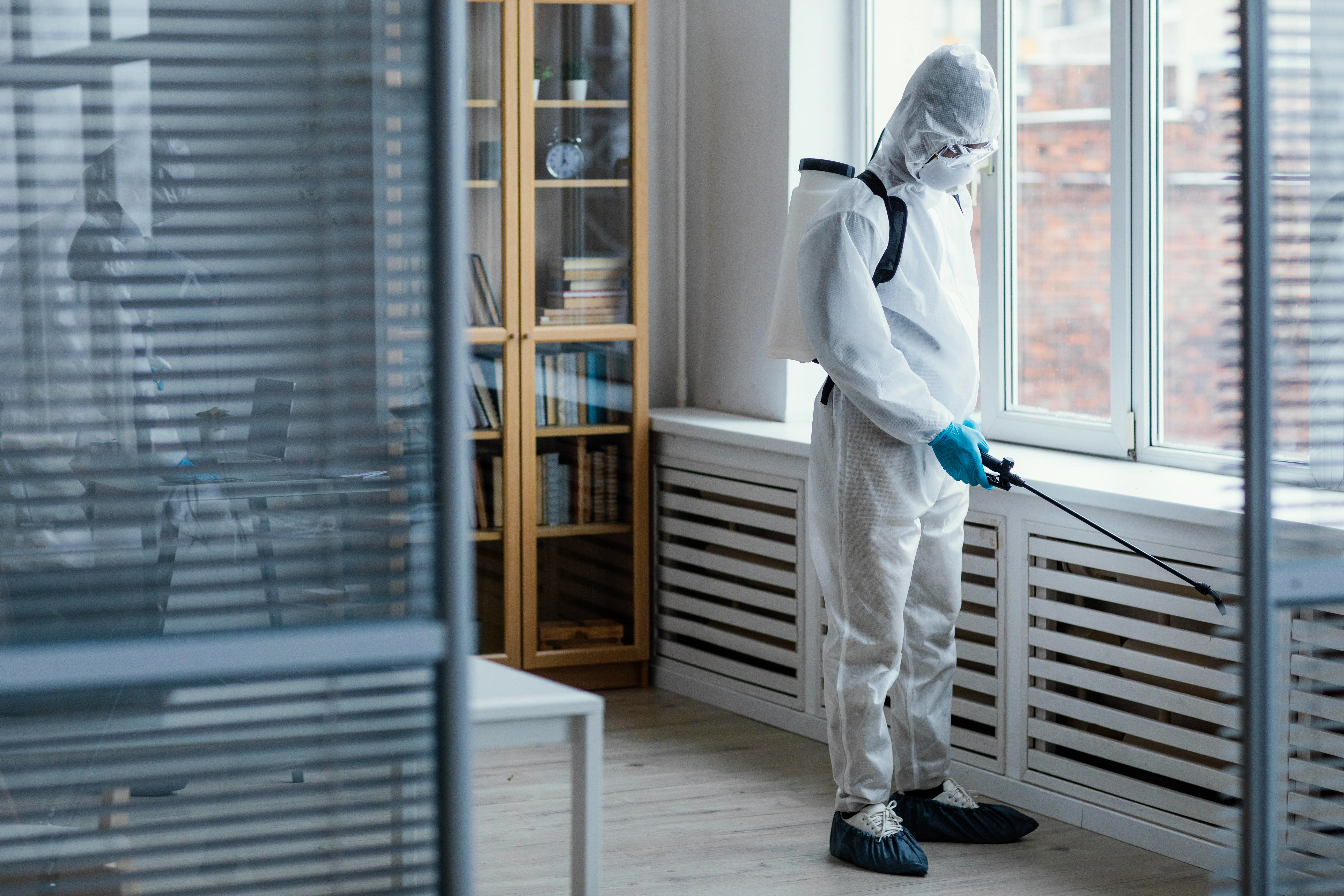 A person in a white protective suit and blue gloves sprays disinfectant near a light office window.