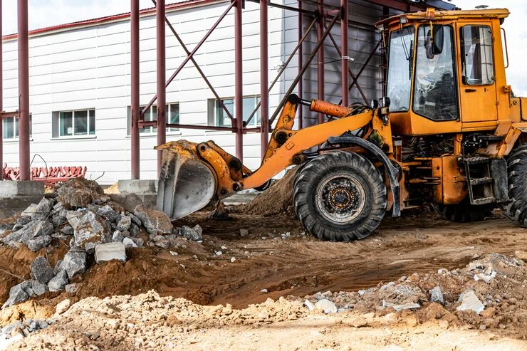 An orange front-loader excavator with its bucket near a pile of concrete debris at a construction site.