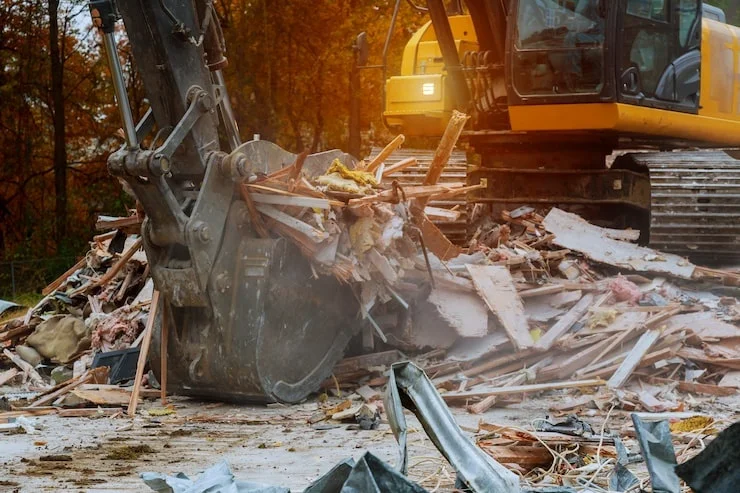 A close-up of a large excavator bucket filled with wood and debris at a demolition site.